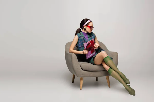 Junge Frau mit Sonnenbrille und Stirnband hält Magazin in der Hand, während sie im Sessel auf grau sitzt — Stockfoto