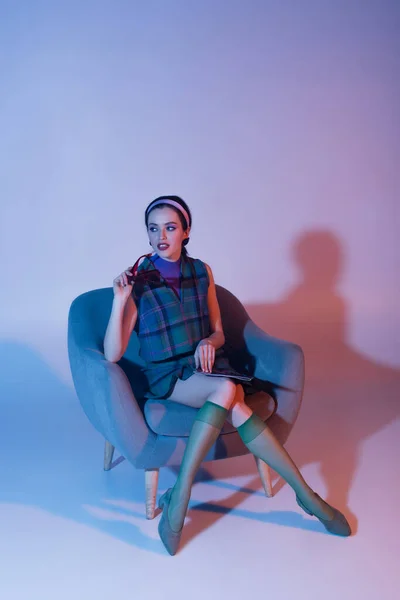 Nachdenkliche junge Frau mit Sonnenbrille und Magazin im Sessel auf lila — Stockfoto