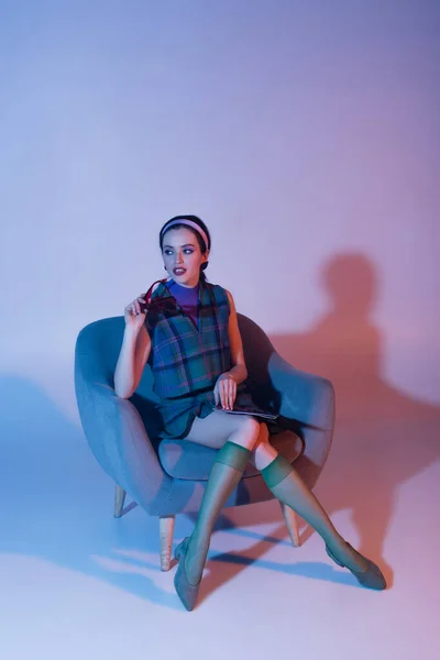 Pensive young woman holding sunglasses and magazine while sitting in armchair on purple — Stock Photo