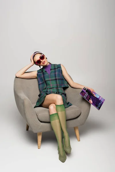 Молодая женщина в солнцезащитных очках держит журнал, сидя в кресле на сером — стоковое фото