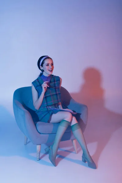 Позитивна молода жінка тримає сонцезахисні окуляри і журнал, сидячи в кріслі на фіолетовому — стокове фото