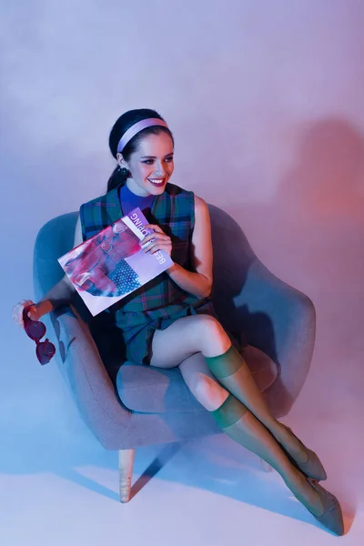 Smiling young woman holding sunglasses and magazine while sitting in armchair on violet — Stock Photo