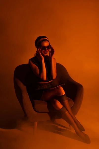 Felice giovane donna che regola gli occhiali da sole e seduta in poltrona con rivista su arancione — Foto stock