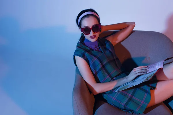 Junge Frau in Sonnenbrille und Retro-Outfit sitzt im Sessel mit Magazin auf blau — Stockfoto