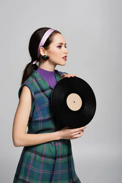 Young brunette woman in headband holding retro vinyl disc isolated on grey — Stock Photo