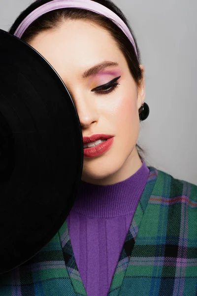 Junge Frau mit Stirnband schaut in der Nähe von Retro-Vinyl-Disc isoliert auf grau — Stockfoto