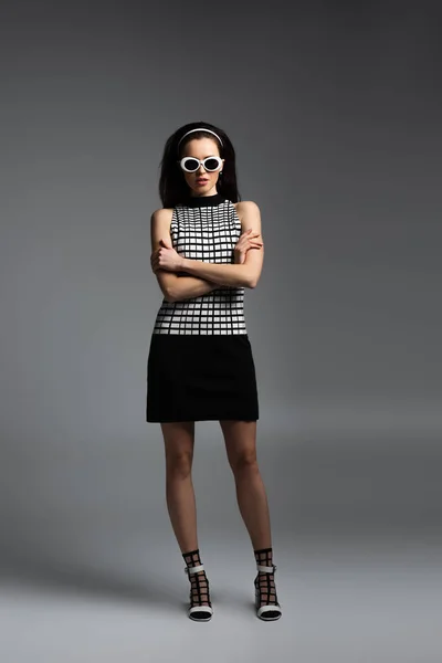 Full length of young brunette woman in sunglasses posing with crossed arms on grey — Stock Photo