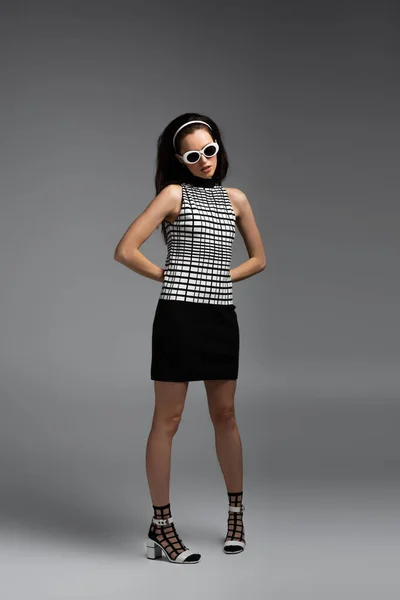 Full length of young brunette woman in sunglasses and retro outfit posing on grey — Stock Photo