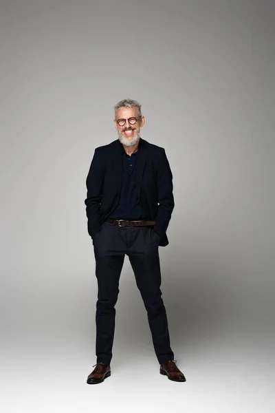 Pleine longueur de joyeux homme d'affaires d'âge moyen dans des lunettes debout avec les mains dans les poches sur le gris — Photo de stock