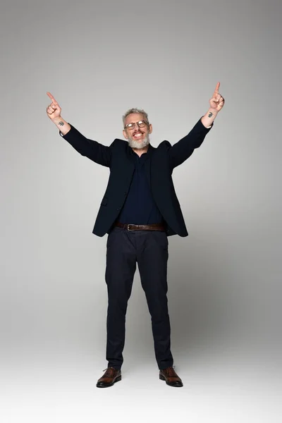 Full length of smiling middle aged man in glasses and suit pointing with fingers on grey — Stock Photo