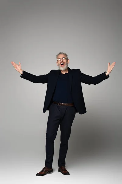 Full length of amazed middle aged man in glasses and suit standing with outstretched hands and singing on grey — Stock Photo