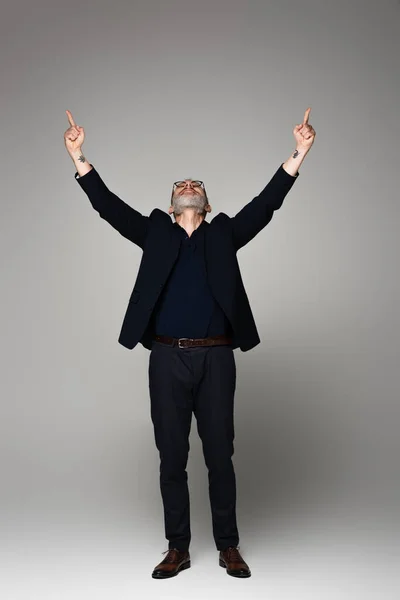 Longitud completa del hombre de mediana edad en gafas y traje apuntando hacia arriba con los dedos en gris - foto de stock