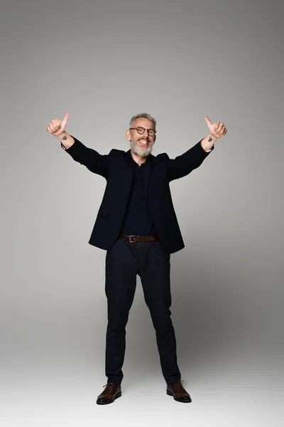 Full length of positive middle aged man in glasses and suit showing thumbs up on grey — Stock Photo