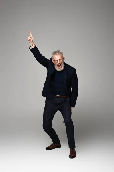 Longitud completa del hombre de mediana edad en gafas y traje gritando mientras señala con el dedo en gris - foto de stock