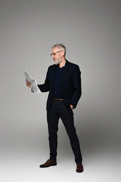 Повнометражний чоловік середнього віку в окулярах і костюмі читає газету, позуючи на сірому — стокове фото