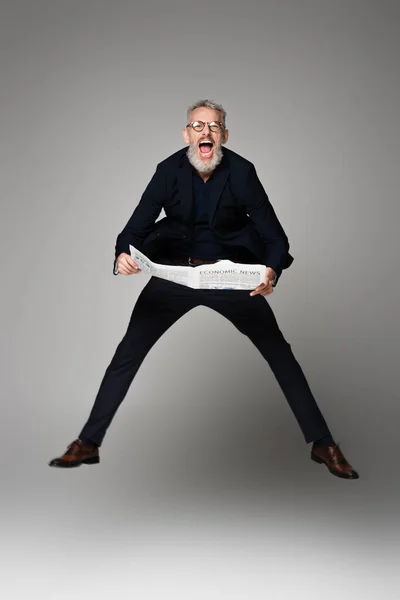 Full length of amazed man in glasses and suit holding newspaper while levitating on grey — Stock Photo