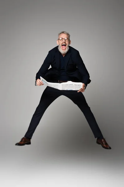 Longitud completa de hombre sorprendido en gafas y traje celebración de periódico mientras levita en gris - foto de stock