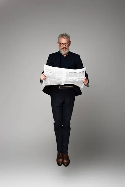 Full length of middle aged businessman in glasses and suit reading economic news while levitating on grey — Stock Photo
