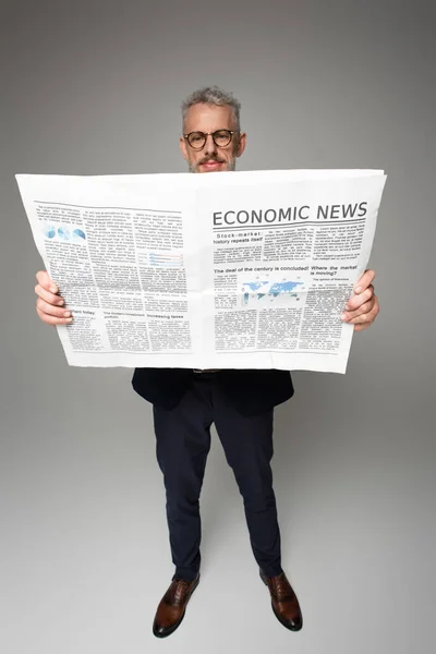 Full length of middle aged businessman in glasses reading economic news on grey — Stock Photo