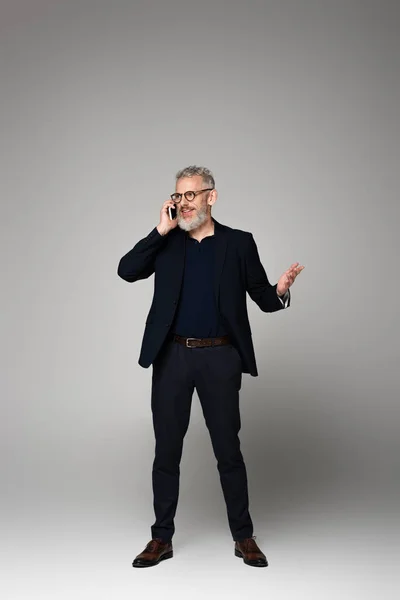 Longitud completa de hombre feliz con el pelo gris hablando en el teléfono inteligente, mientras que el gesto en gris - foto de stock