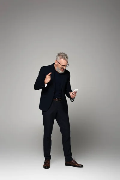 Full length of happy businessman with grey hair using smartphone while pointing with finger on grey — Stock Photo