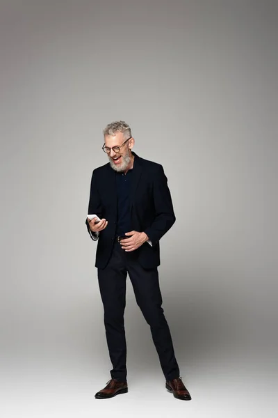 Comprimento total de empresário animado em terno usando smartphone em cinza — Fotografia de Stock