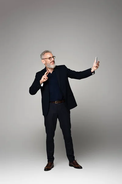 Glücklicher Geschäftsmann mit grauen Haaren, der ein Selfie mit dem Smartphone macht, während er ein Friedenszeichen auf Grau zeigt — Stockfoto
