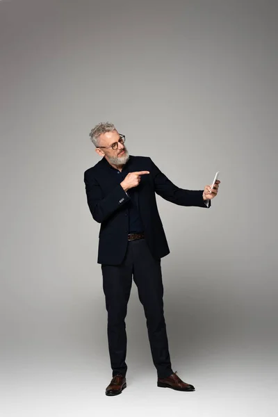 Full length of businessman with grey hair pointing with finger while taking selfie on grey — Stock Photo