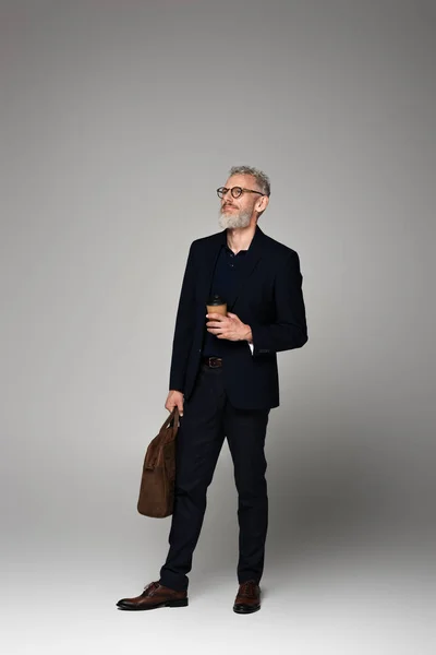 Full length of man with grey hair in suit holding paper cup and leather bag on grey — Stock Photo