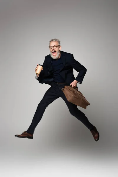 Longitud completa del hombre excitado con pelo gris en traje sosteniendo taza de papel y bolsa de cuero mientras levita sobre gris - foto de stock