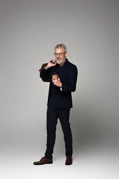 Full length of man with grey hair holding paper cup and leather bag while standing on grey — Stock Photo