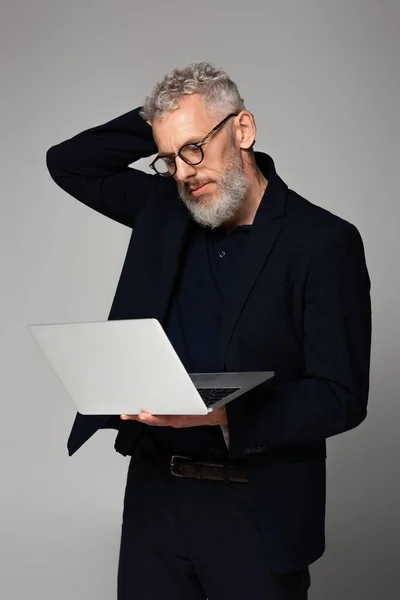Pensoso uomo di mezza età in occhiali guardando computer portatile isolato su grigio — Foto stock