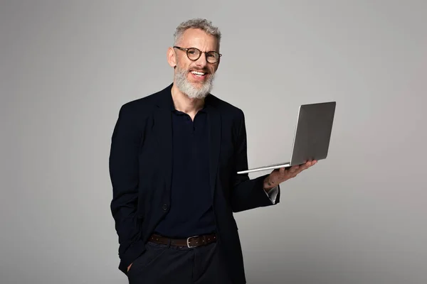 Felice uomo con i capelli grigi che tiene il computer portatile mentre in piedi con mano in tasca isolato su grigio — Foto stock