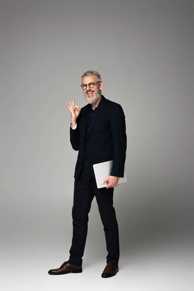 Full length of happy man with grey hair holding laptop while showing ok sign on grey — Stock Photo