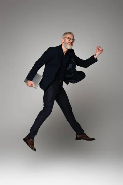 Full length of happy man with grey hair holding laptop while levitating on grey — Stock Photo