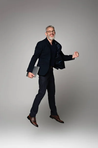 Full length of cheerful man with grey hair holding laptop while levitating on grey — Stock Photo