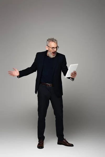 Comprimento total de homem de negócios irritado de meia idade com cabelos grisalhos gritando enquanto olha para tablet digital em cinza — Fotografia de Stock