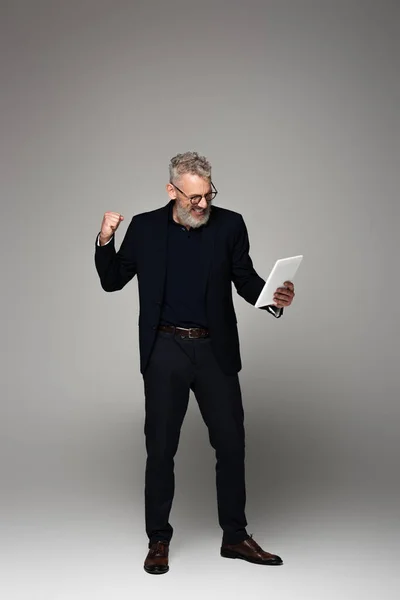 Pleine longueur d'homme d'affaires excité d'âge moyen avec les cheveux gris regardant la tablette numérique et se réjouissant sur gris — Photo de stock