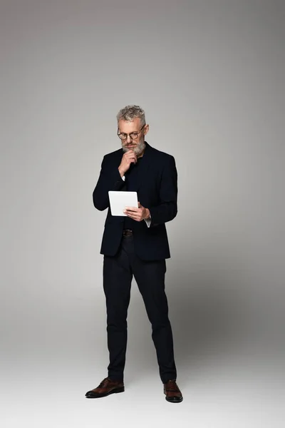 Volle Länge des nachdenklichen Mannes mittleren Alters mit grauen Haaren, der das digitale Tablet auf grau betrachtet — Stockfoto