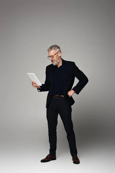 Full length of man with grey hair looking at digital tablet and posing on grey — Stock Photo