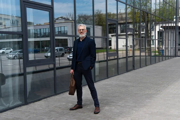 Full length of mature man with grey hair holding leather bag while standing outside — Stock Photo