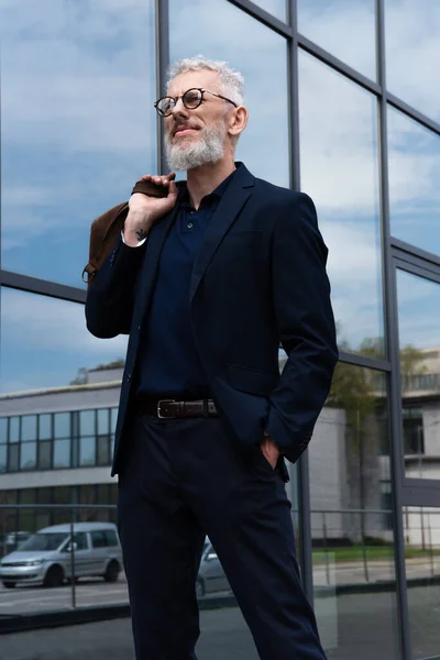 Tattooed mature man with grey hair holding leather bag while standing outside — Stock Photo