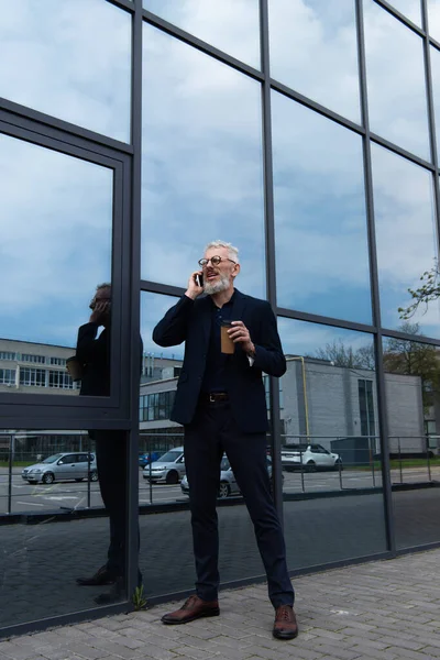 Piena lunghezza dell'uomo maturo con i capelli grigi in possesso di tazza di carta mentre parla su smartphone vicino all'edificio moderno — Foto stock