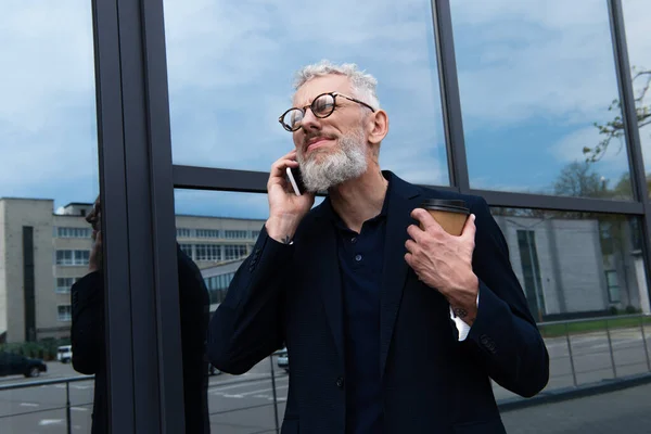 Hombre de negocios con pelo gris sosteniendo una taza de papel mientras habla en un teléfono inteligente cerca de un edificio moderno - foto de stock