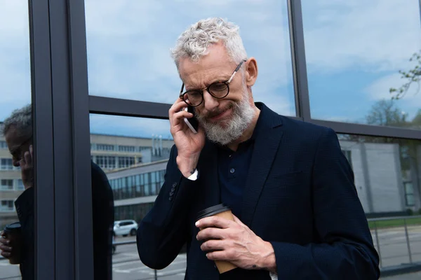 Confuso hombre de negocios con pelo gris sosteniendo una taza de papel mientras habla en un teléfono inteligente cerca de un edificio moderno - foto de stock