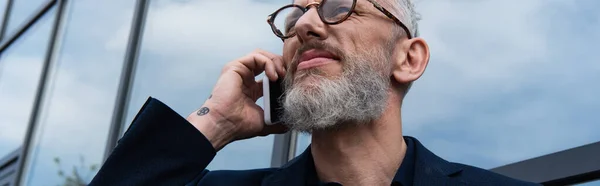Зрілий бізнесмен з сірим волоссям розмовляє на смартфоні біля сучасної будівлі, банер — стокове фото