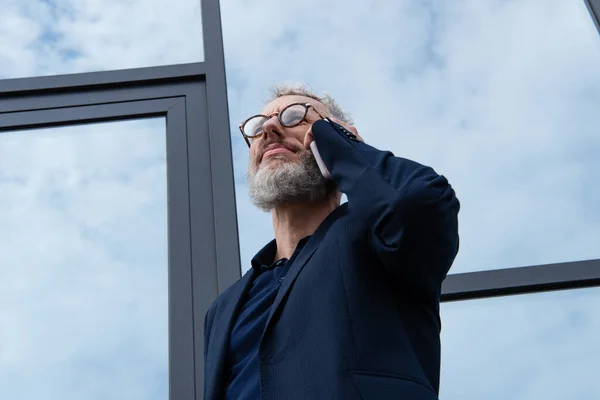 Reifer Geschäftsmann mit Brille und grauen Haaren spricht auf dem Smartphone in der Nähe moderner Gebäude — Stockfoto