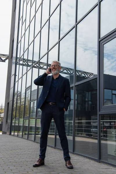 Volle Länge des glücklichen Geschäftsmannes mittleren Alters in Brille, der auf dem Smartphone in der Nähe des Gebäudes spricht — Stockfoto