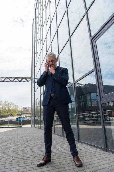 Full length of pensive middle aged businessman in glasses talking on smartphone near building — Stock Photo