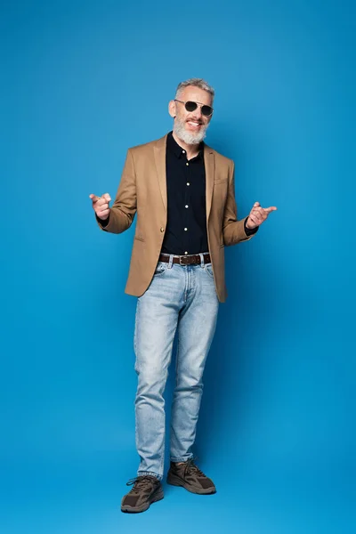 Pleine longueur de heureux homme d'âge moyen dans des lunettes de soleil pointant avec les doigts sur le bleu — Photo de stock
